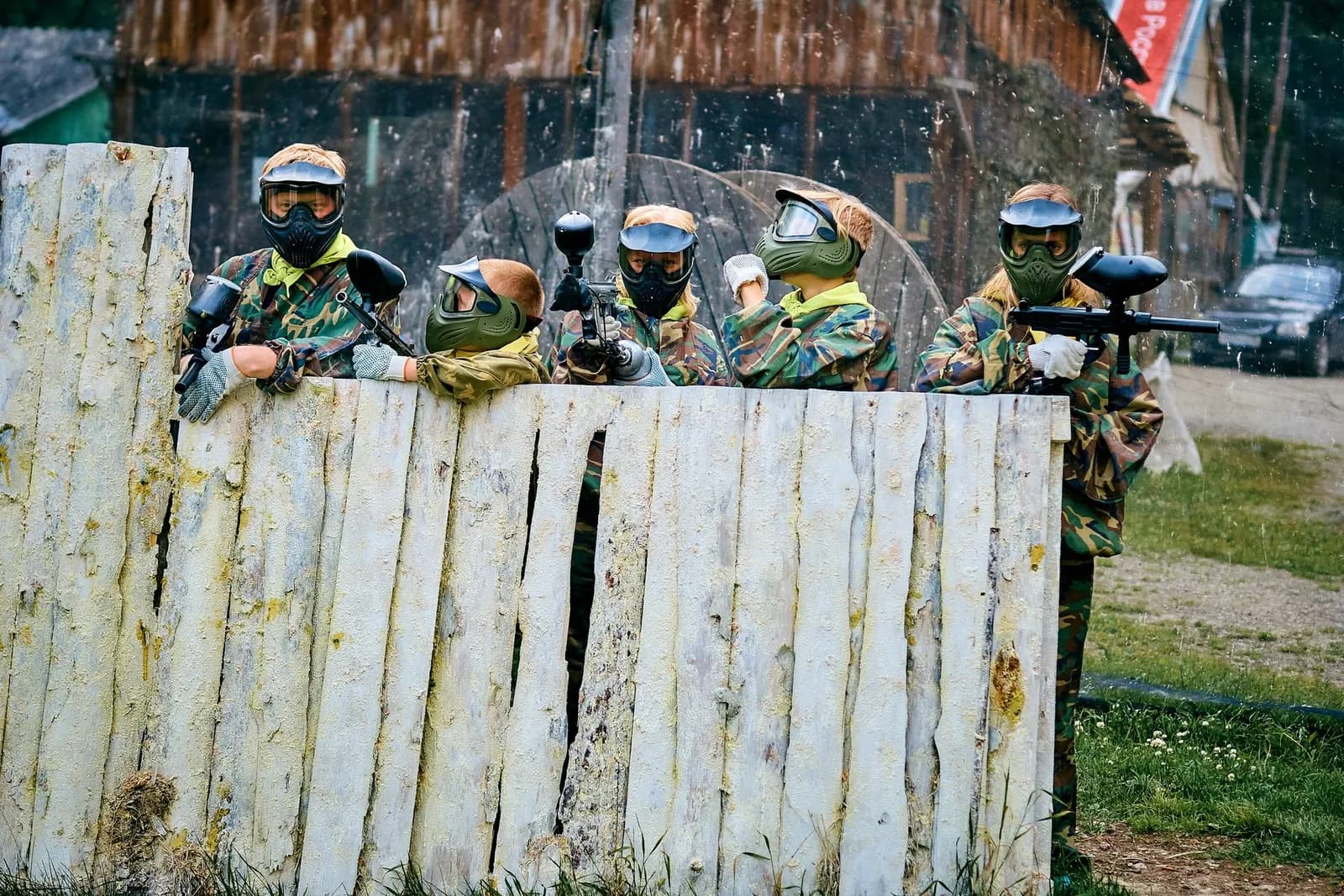 Équipe de joueurs de paintball masqués alignés derrière une palissade en bois, lanceurs prêts