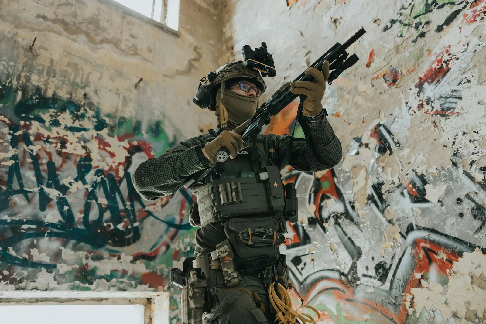 Joueur d'airsoft équipé, réplique levée, progressant dans un bâtiment aux murs couverts de graffitis