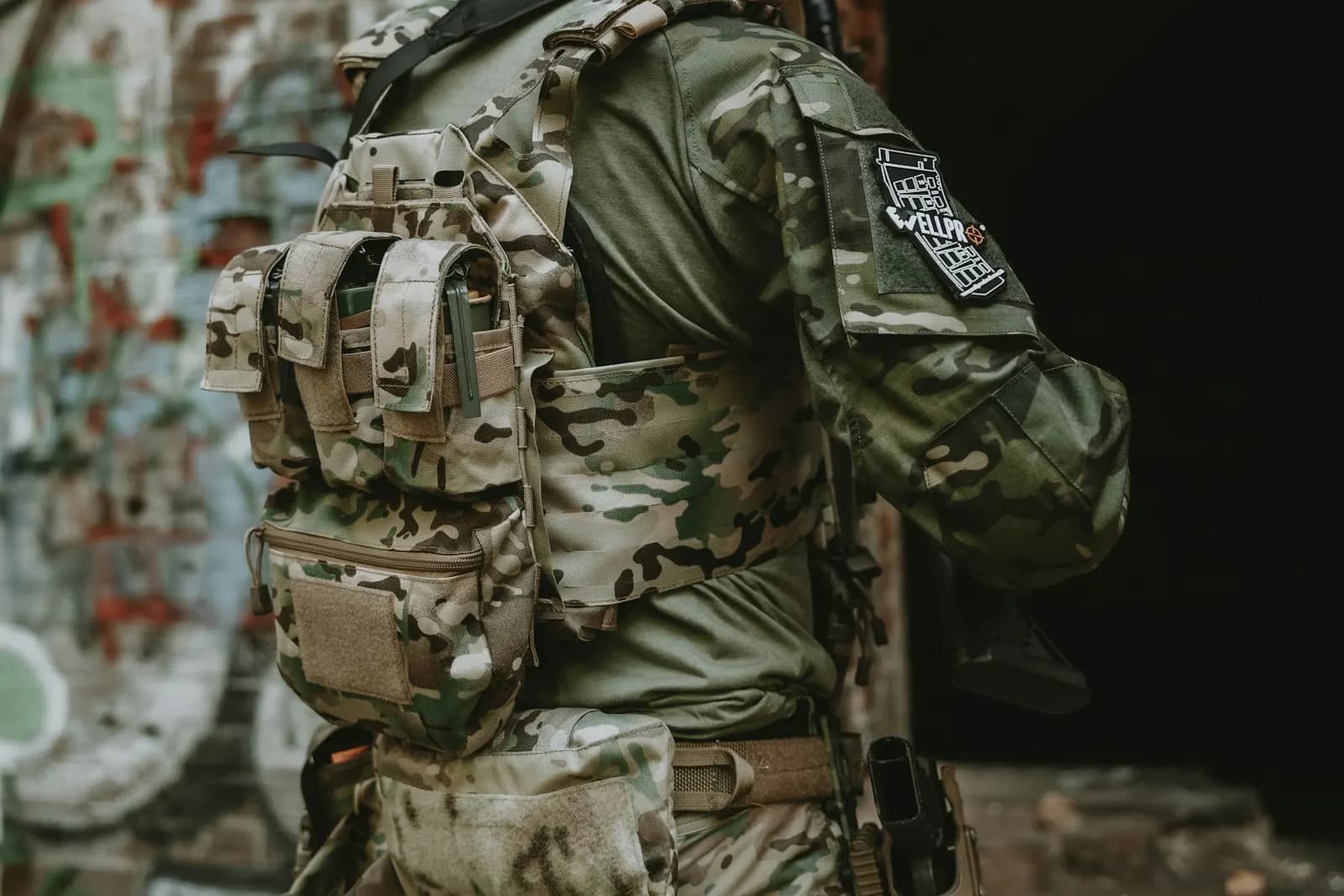 Joueur d'airsoft vu de dos portant un gilet porte-plaques camouflage équipé de poches, dans un bâtiment