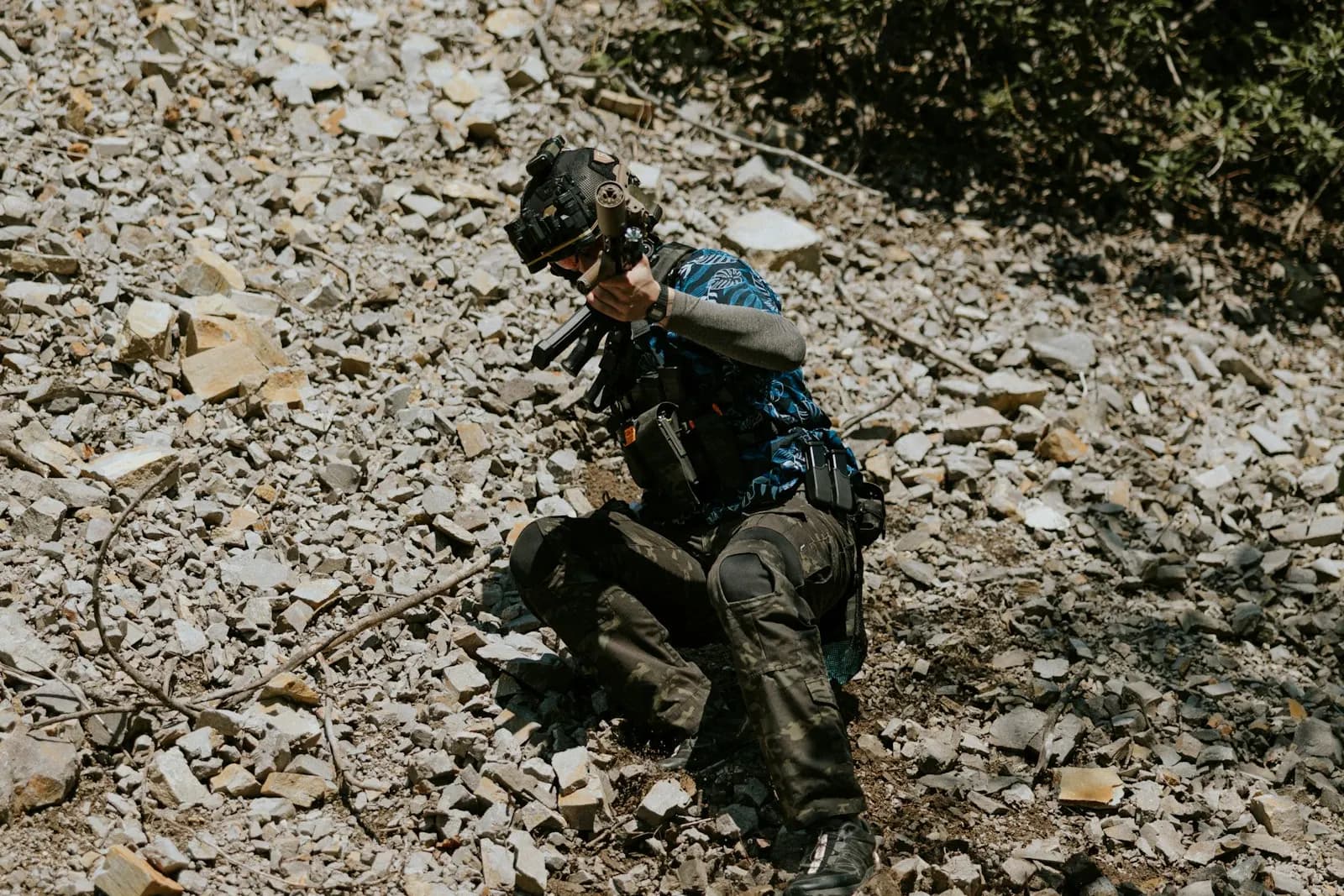 Joueur d'airsoft accroupi sur un terrain rocailleux, équipé et réplique en main