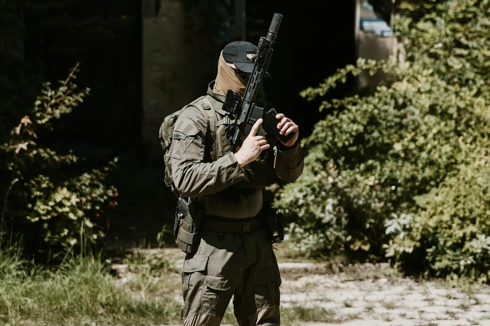 Joueur d'airsoft en tenue tactique olive, cagoule et casquette, tenant sa réplique dans un terrain arboré