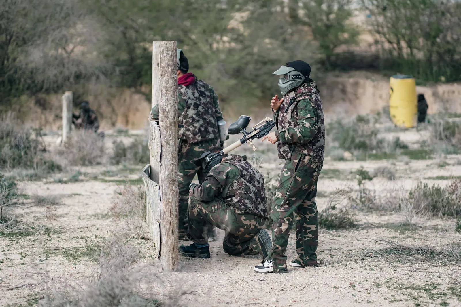 Joueurs de paintball en tenue camouflage derrière un poteau en bois sur un terrain sablonneux
