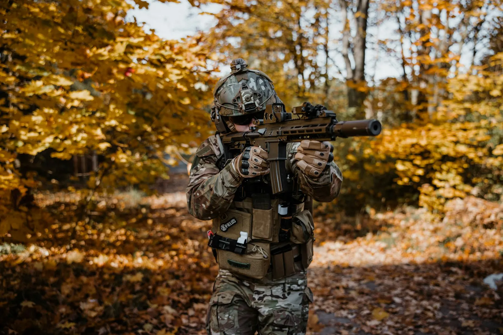 Joueur d'airsoft équipé, réplique épaulée, progressant sur un chemin couvert de feuilles mortes dans une forêt d'automne