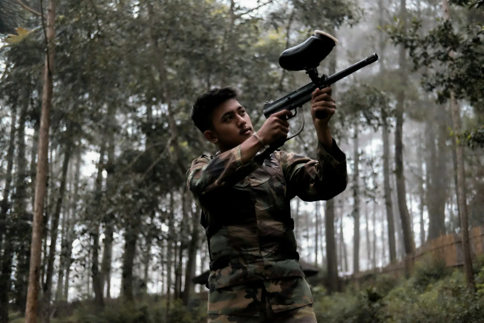 Joueur de paintball en tenue camouflage visant avec un lanceur équipé d'une trémie dans une forêt de pins