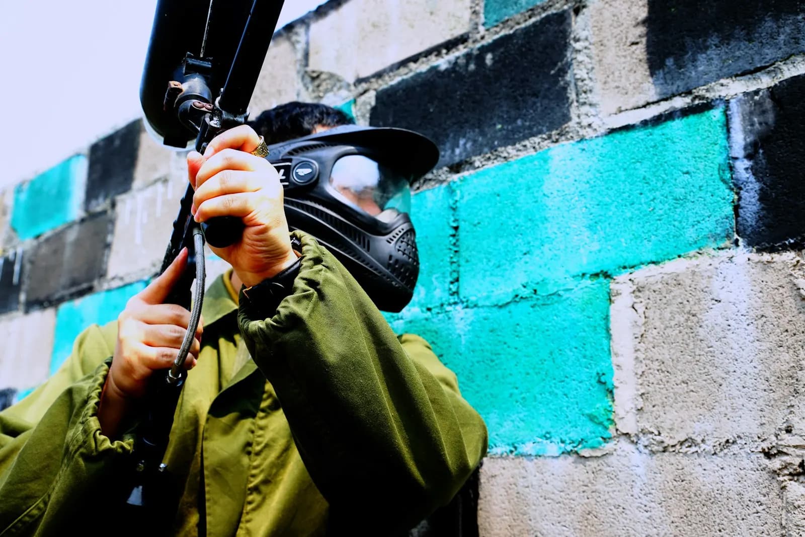 Joueur de paintball portant un masque et une veste verte, lanceur levé, à l'abri contre un mur de parpaings colorés