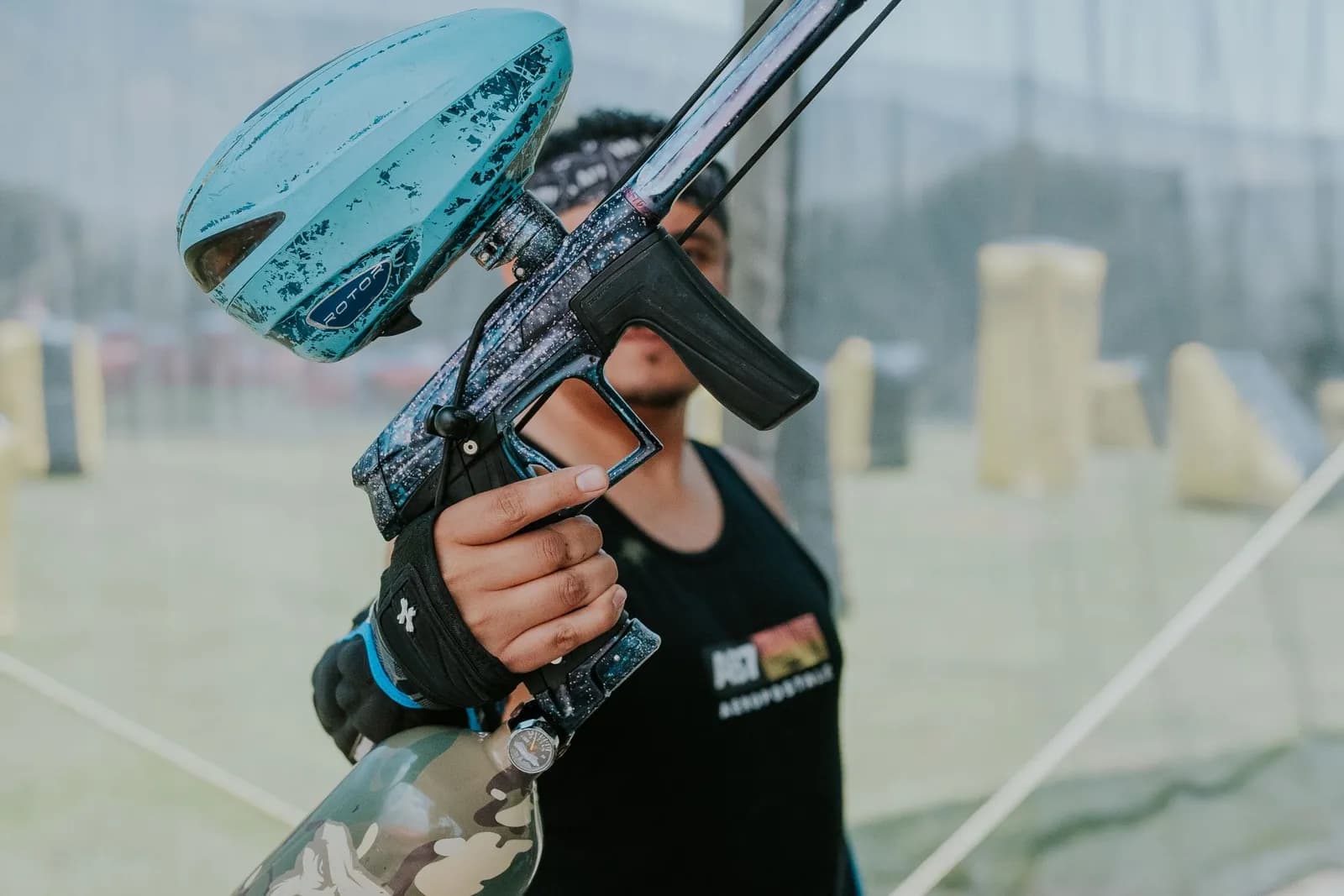 Joueur de paintball tenant un lanceur à trémie bleue avec un canon décoré, sur un terrain