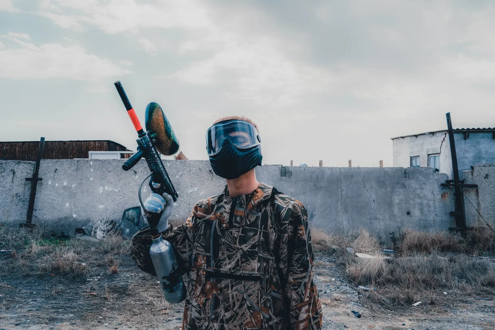 Joueur de paintball en tenue camouflage tenant un lanceur mécanique avec trémie devant un mur