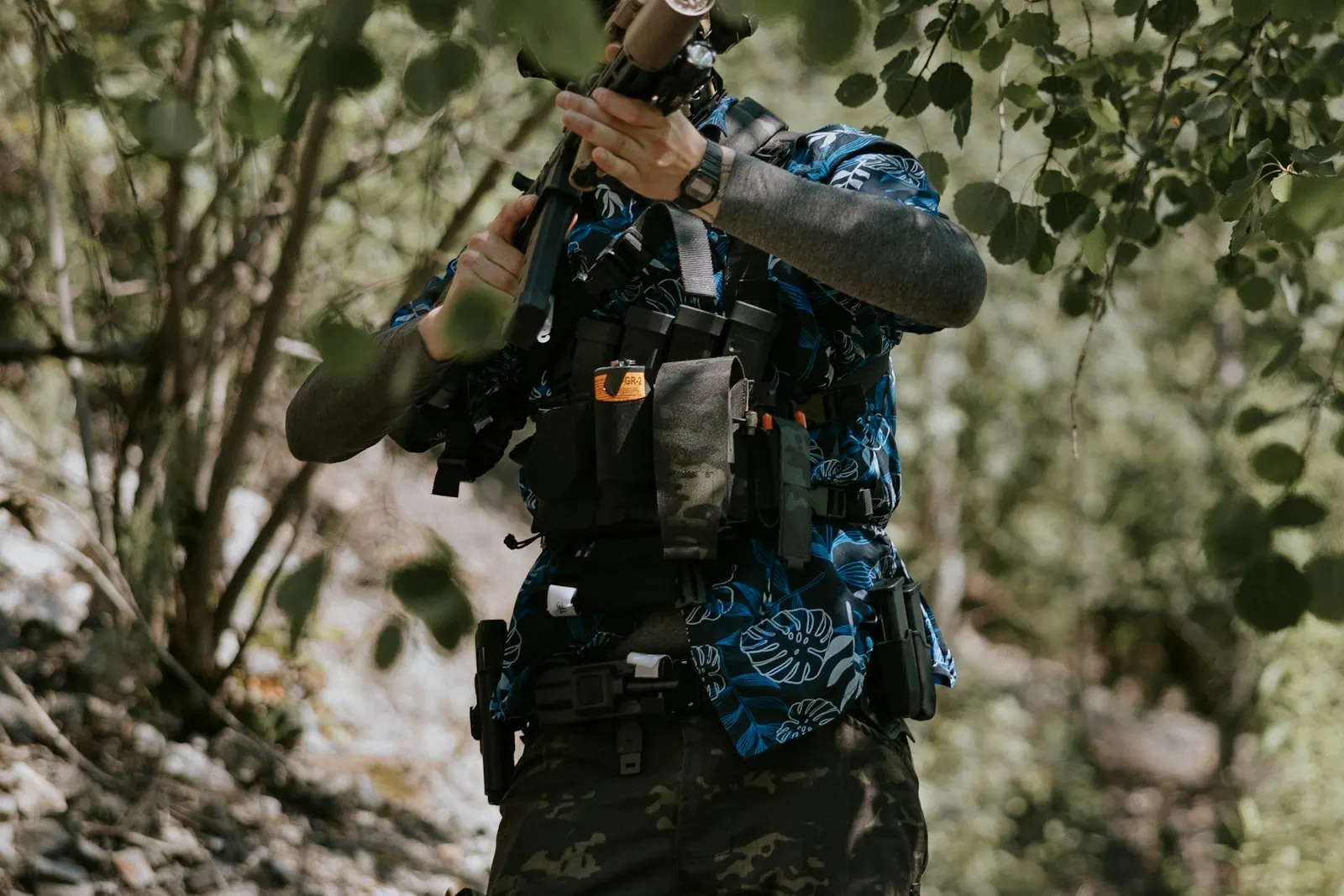 Joueur d'airsoft en chemise à motifs et gilet tactique, réplique épaulée, progressant en sous-bois