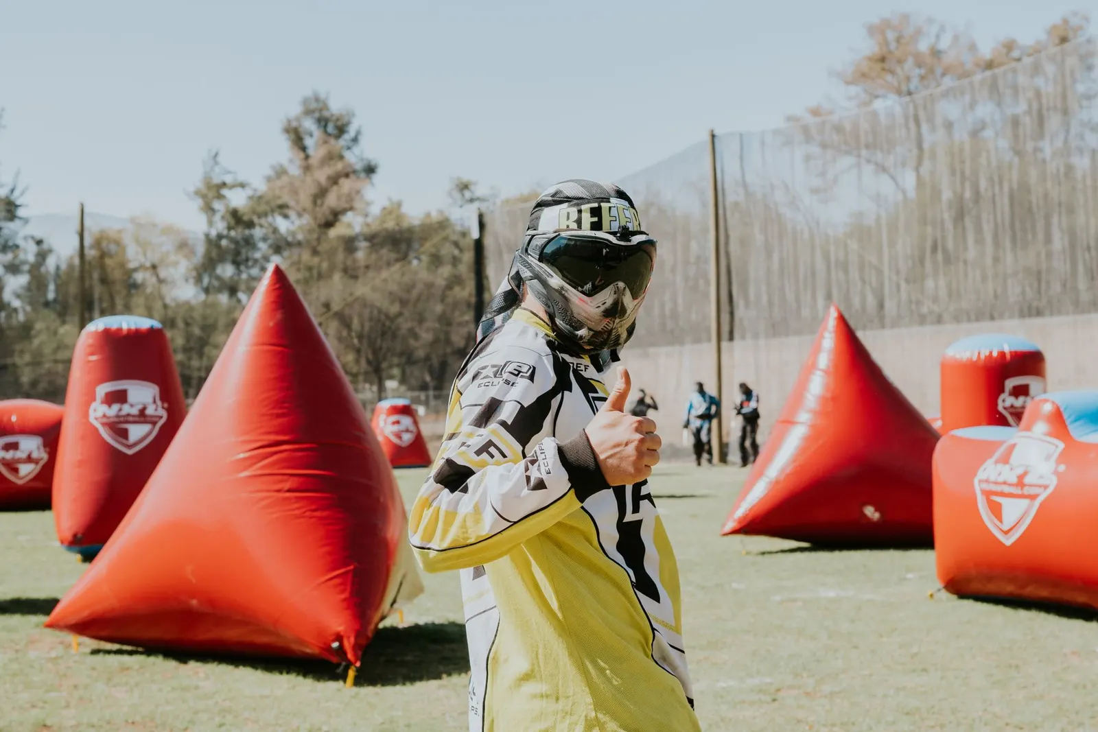 Joueur de paintball en tenue jaune et masque faisant un pouce levé sur un terrain d'obstacles gonflables