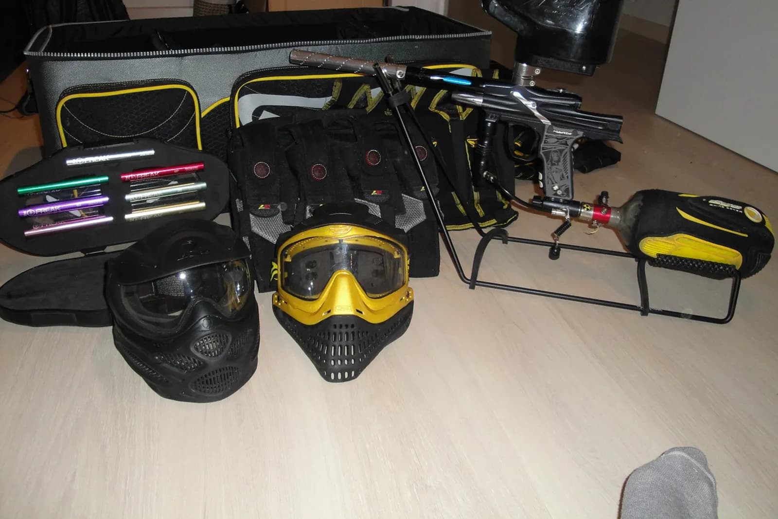Équipement de paintball posé au sol : masques, lanceur, sac et tubes de billes colorés