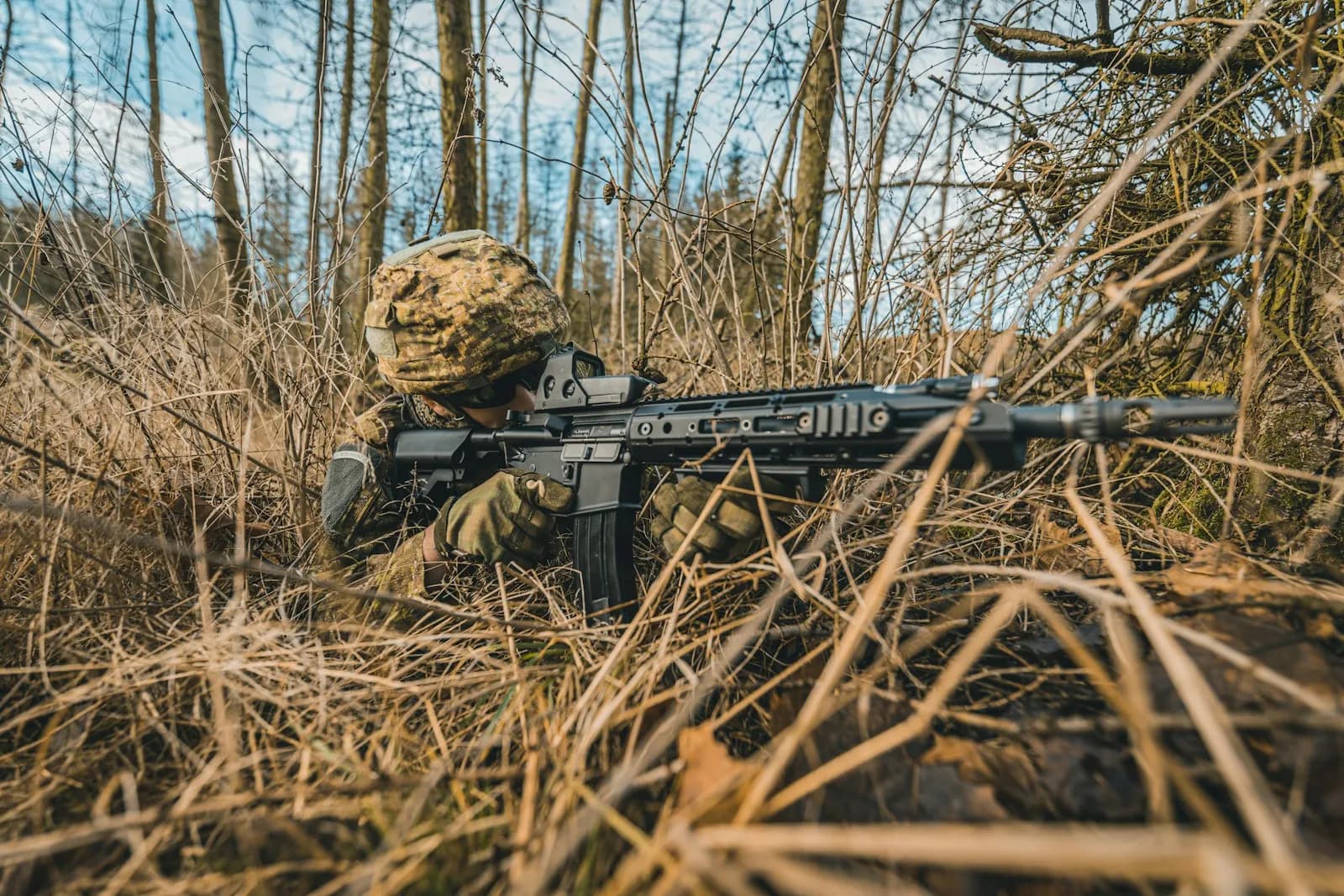 Joueur d'airsoft allongé dans les herbes hautes d'une forêt, en position de tir avec sa réplique