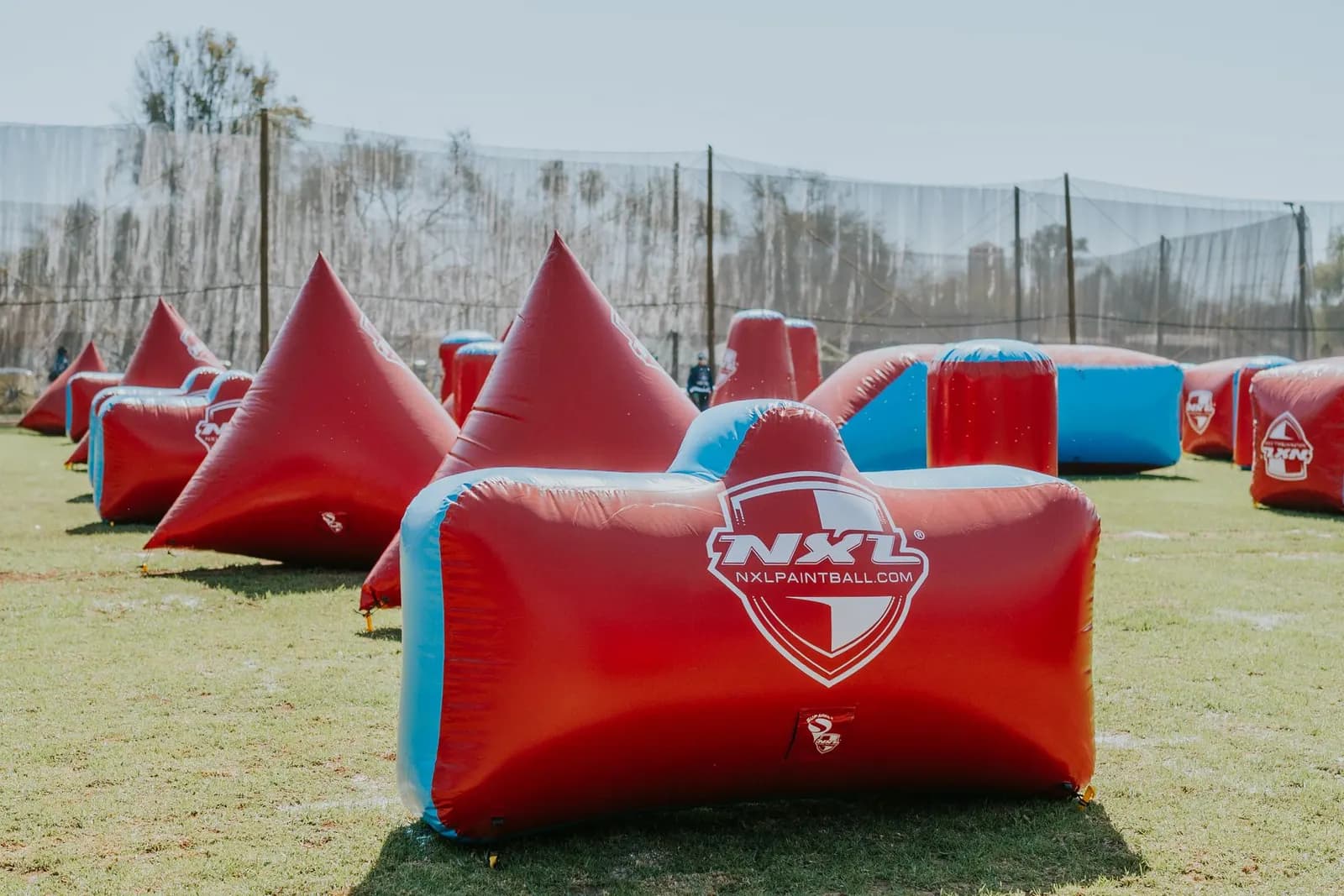 Terrain de speedball avec obstacles gonflables rouges et bleus en forme de pyramides, cylindres et boudins, entouré de filets