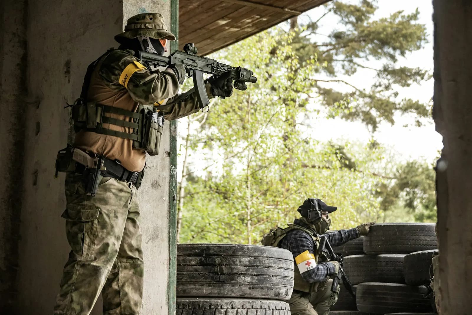 Joueur d'airsoft en tenue tactique visant depuis l'angle d'un bâtiment, un autre joueur à couvert derrière des pneus