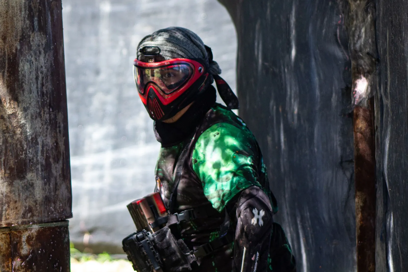 Joueur de paintball en tenue complète, masque rouge et maillot vert, derrière un obstacle