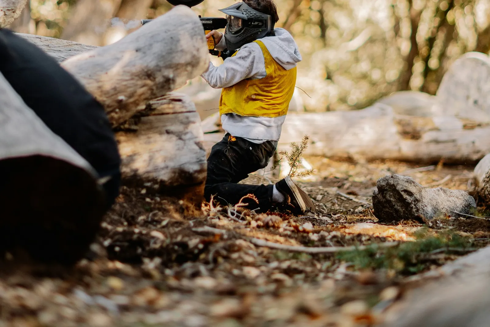 Joueur de paintball à genou derrière des troncs couchés dans une forêt, masque sur le visage