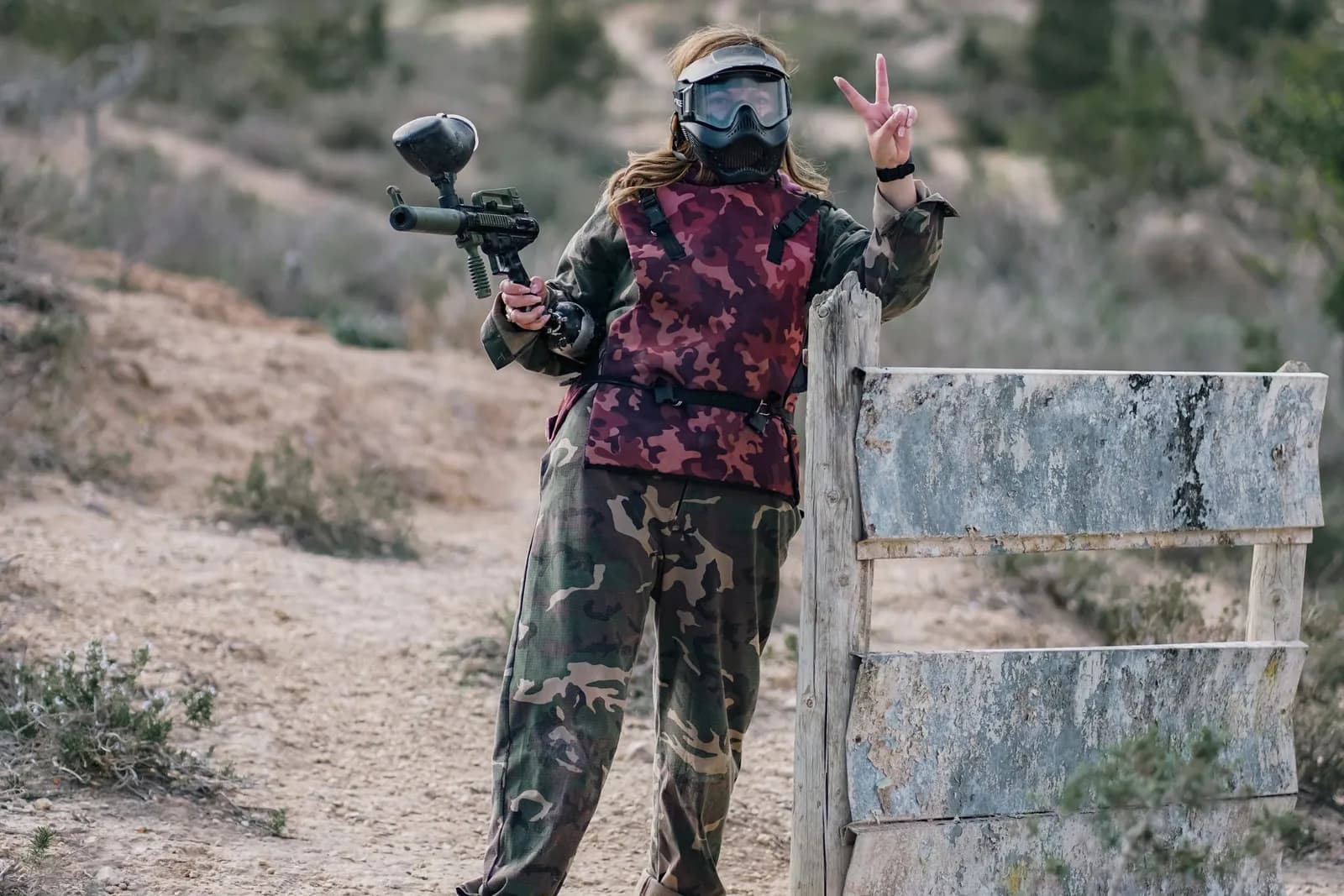 Joueuse de paintball en tenue camouflage et masque sur un terrain naturel vallonné près d'une palissade